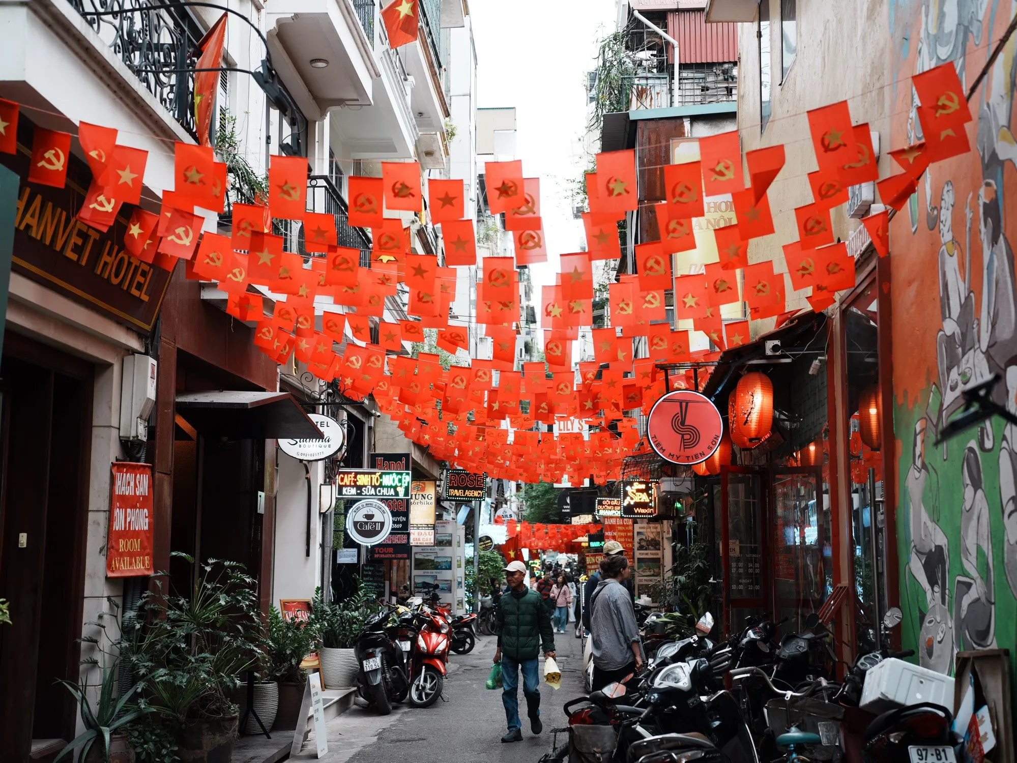 A street in Hanoi lined with numerous red Vietnamese national flags and red party flags hanging closely together across buildings and sidewalks.